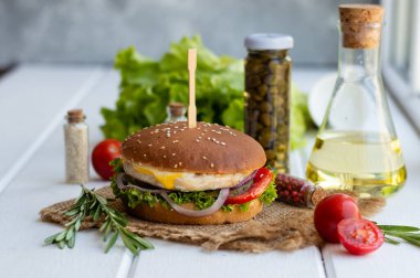 Kızarmış patatesli tavuk burger, kiraz domatesli kapari, salata yaprakları, sos ve biberiye, beyaz ahşap bir yüzey üzerinde..