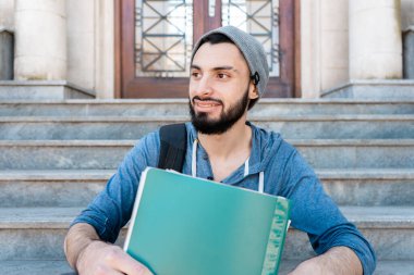 Sırt çantası ve klasörlü öğrenci üniversite merdivenlerinde oturuyor, yan tarafa bakıyor ve gülümsüyor.