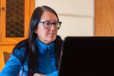 Orta yaşlı bir kadın laptopuyla evden çalışıyor, mavi giyiyor ve gözlük takıyor..