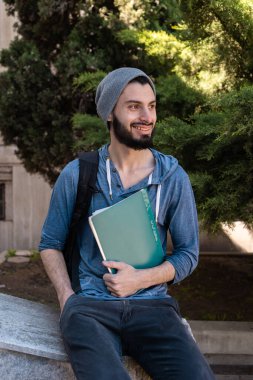 Dikey çekim. Sırt çantası ve kitapları olan bir öğrenci yan bakıp gülüyor. Günlük kıyafet ve bitki örtüsü.