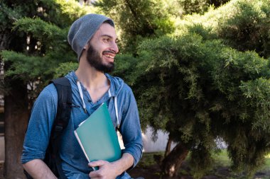 Sırt çantalı ve kitaplı öğrenci yan tarafa bakıyor ve günlük kıyafet ve bitki örtüsü arka planında gülüyor..