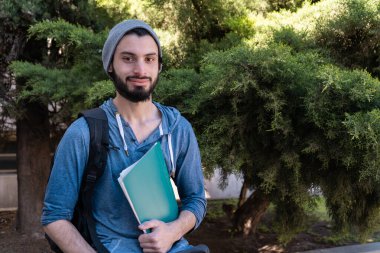 Sırt çantalı ve kitaplı bir öğrenci hafif bir gülümsemeyle kameraya bakıyor. Günlük kıyafet ve bitki örtüsü..