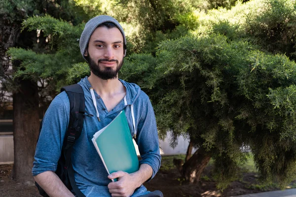 Sırt çantalı ve kitaplı bir öğrenci hafif bir gülümsemeyle kameraya bakıyor. Günlük kıyafet ve bitki örtüsü..