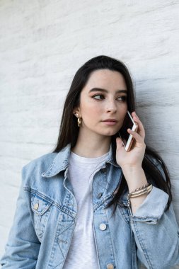 Beyaz bir duvara yaslanmış, telefonda konuşan siyah saçlı genç bir kadın. Dikey çekim. 