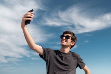Düşük açılı çekimde, arka planda dramatik mavi bir gökyüzü olan genç bir adam selfie çekip kameraya gülümsüyor.