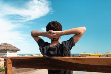 Genç adam bir bankta oturuyor ve plajda yaz tatilinin tadını çıkarıyor..