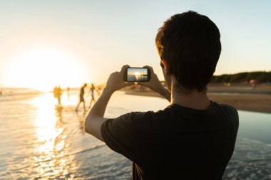 Gün batımında güneşin fotoğrafını çeken genç bir adam..