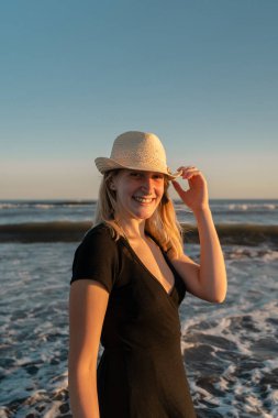 Gün batımında sahilde genç bir kadın kameraya gülüyor ve gülümsüyor. Dikey çekim.