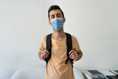 Evde genç bir öğrenci kameraya bakıyor ve maske ve sırt çantası takıyor.. 