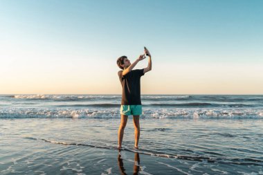 Gün batımında sahilde genç bir adam akıllı telefonunda 5G internet sinyali arıyor..