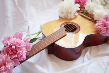An acoustic wooden guitar lies on a white sheet surrounded by pink and white peonies. The concept of learning music, romantic serenade.