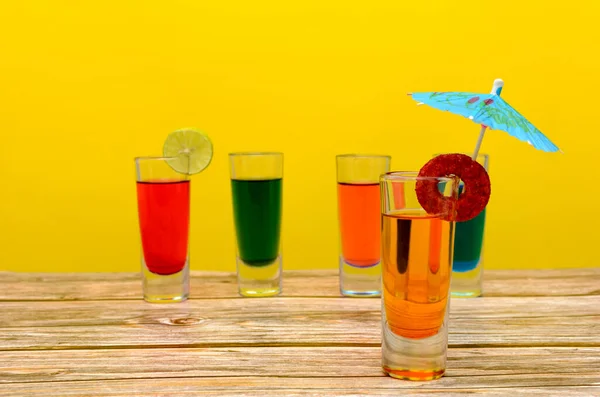 Colorful drinks on wooden table. Small glass glasses with colorful drinks on wooden table with yellow background.