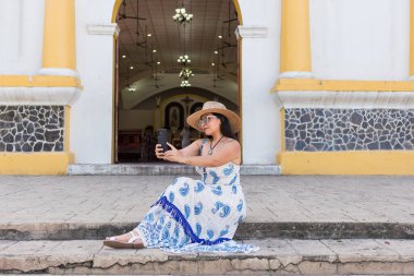 Şapkalı, mavi beyaz elbiseli bir kadın bir kilisenin merdivenlerine oturur ve selfie çeker..