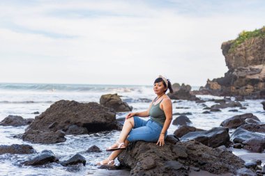 Dalgın bir kadın sahildeki büyük bir kayanın üzerinde huzur içinde oturur ve okyanusa bakar.