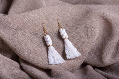 Bir çift beyaz makrome püskül küpe altın aksanlı desenli bej kumaş üzerinde dinleniyor..