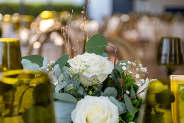 Beyaz güller, okaliptüs yaprakları ve küçük beyaz çiçeklerden oluşan zarif bir masa dekorasyonunun yakın çekimi..