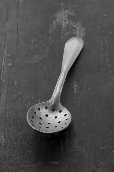 Vintage aluminum spoon with holes on an old battered black table closeup. selective focus. black and white photo. shallow depth of field — Stock Photo, Image