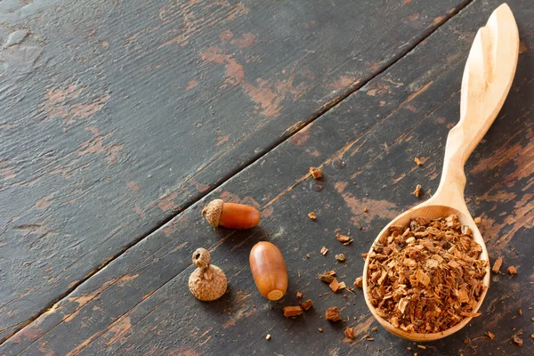 Bellotas y corteza de roble triturada en una cuchara de madera sobre una vieja mesa de madera negra. materias primas vegetales para la preparación de bebidas de té, medicamento. medicina alternativa. Copiar espacio. Espacio libre para texto. enfoque selectivo — Foto de Stock