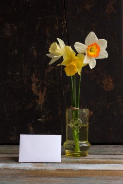 Three spring flower yellow and white daffodils with Golden wedding rings in glass vase with greeting blank card on old wooden background black. The Provence style, rustic. With space for text