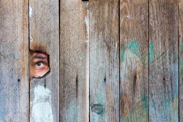 The eyes of a man spying through a hole in an old wooden fence — Stock Photo, Image