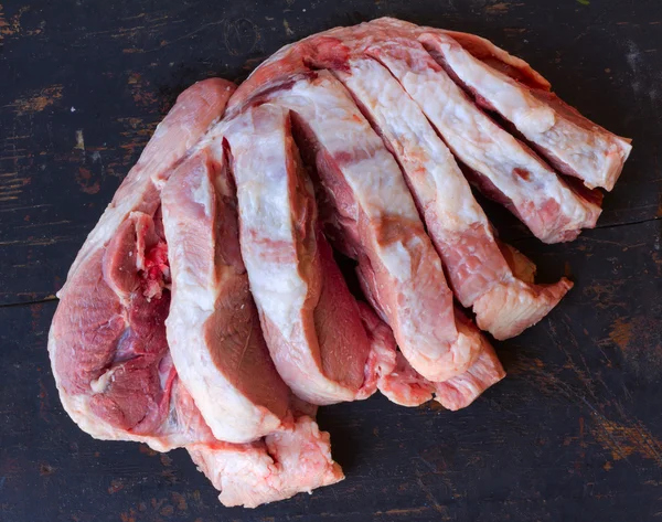 Egy nagy darab nyers sertéshús vágott darabokra, a régi fekete vágódeszka, közeli felülnézet — Stock Fotó