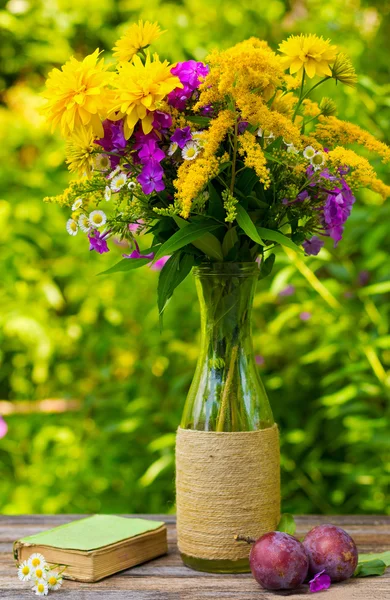 Un bouquet de fleurs de verge d'or, phlox chrysanthèmes dans un vase en verre, prunes mûres et vieux livre avec marguerites sur la table sur fond de nature — Photo