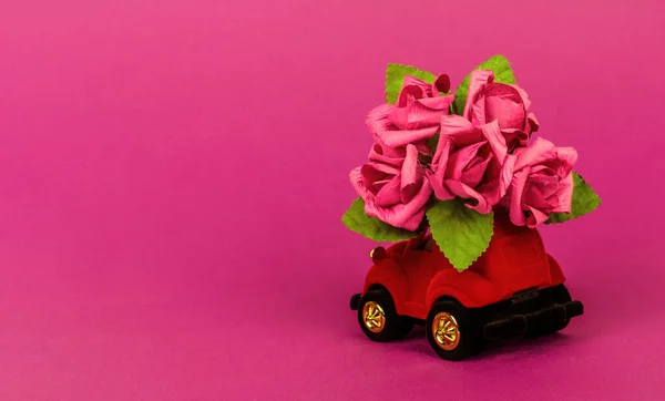 red velour toy car delivering bouquet of flowers on pink background with copy space. Valentine\'s day card, birthday, 8 March, International Happy Womens Day