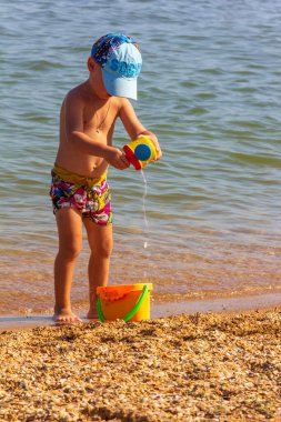Oyuncak kovası ve su dolu tenekesi olan küçük bir çocuk yaz günü kumsalda oynar.