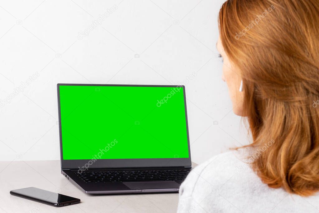 young woman sitting in front of a laptop with a green mockup on the screen, concept of distance learning, live broadcast, online communication