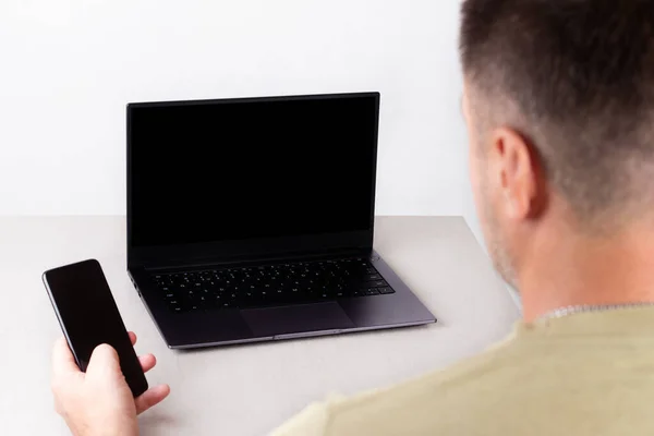 a man with a phone in his hand sits in front of a laptop with a black mock-up on the monitor, the concept of office work, remote work, marketing, training, coaching