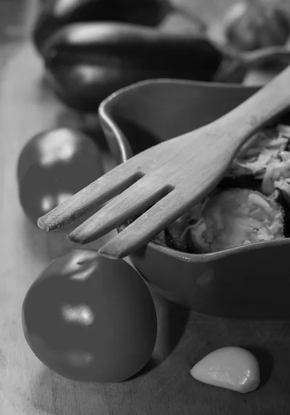 Trägaffel, stekt aubergine med vitlök i en keramikskål och råa vitlöksklyftor, tomat och aubergine i svart och vitt, selektiv fokus — Stockfoto