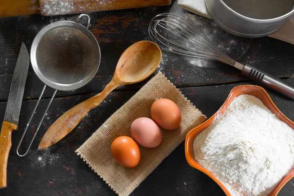 Küchenutensilien und Lebensmittel am alten heimischen Küchentisch. Hühnereier und Weizenmehl für die Teigvorbereitung. rustikaler Stil. Nahaufnahme von oben. — Stockfoto