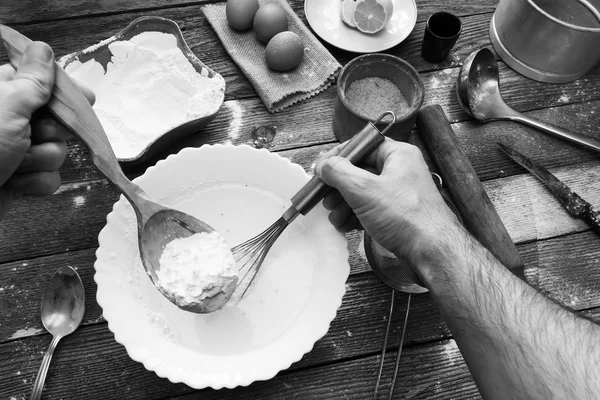 男は、泡立て器で生地をこねます。木製のテーブルに小麦の粉、ねり粉、卵、レモン、キッチン道具。素朴なキッチンで生地の準備。黒と白の写真 — ストック写真