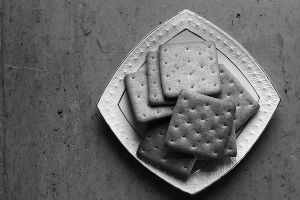 Cracker biscuits in a white ceramic square saucer on old wooden background close-up. Black and white photo. Free space for text. Copy space. Top view — Stock Photo, Image