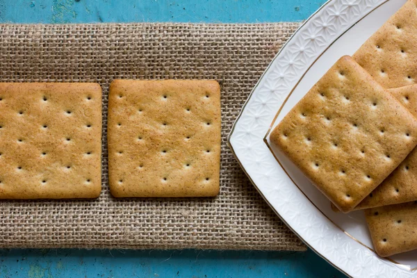 Galletas de galletas cuadradas en una servilleta de lino y una porción triangular de platos de cerámica blanca con galletas en la vieja mesa azul. Primer plano, vista superior —  Fotos de Stock