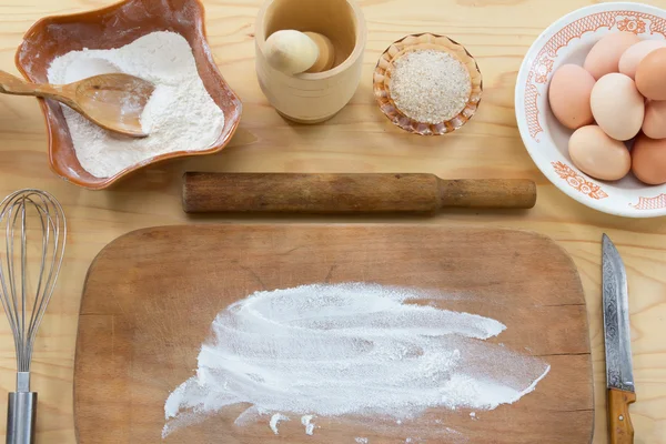 Das alte Nudelholz und ein Holzschneidebrett mit Mehl, Hühnereiern, Schneebesen, Messer und Mörser mit Stößel auf dem Holztisch — Stockfoto