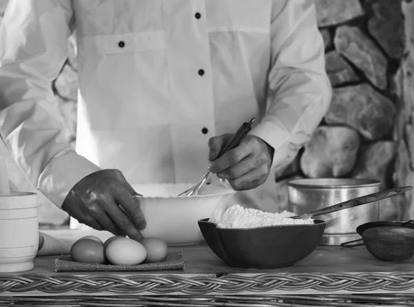 Em primeiro plano uma tigela com farinha, ovos, peneira e argamassa em segundo plano um homem vestido com chicotes brancos batem a massa. conceito de cozinha rústica, foco seletivo. foto preto e branco — Fotografia de Stock