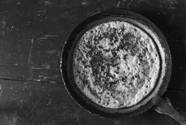 Burnt pancake from wheat flour in an old frying pan on a black background. Rustic style. Top view closeup. Black and white photo — Stock Photo, Image