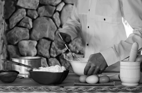 Farinha, ovos, uma peneira e uma argamassa, e um cozinheiro macho em uma peça de roupa branca batem a massa. Conceito de cozinha rústica, foco seletivo. Espaço para cópia. Espaço livre para texto. foto preto e branco — Fotografia de Stock