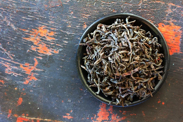 Torkade svart te lämnar i rund svart tefat på gamla trä bakgrund. Kopiera utrymme. Fritt utrymme för text, närbild, ovanifrån — Stockfoto