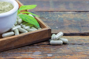 Andrographis panikleme kapsülleri, panikleme tozu ve yapraklar tahta tabakta ve ahşap arka planda.