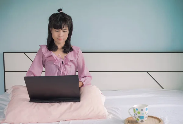 Asian woman sitting on the bed with laptop. She is blogger using taplet for work on social and shopping online and write to friend  by network. her tan skin and black heir authentic. holidays concept.