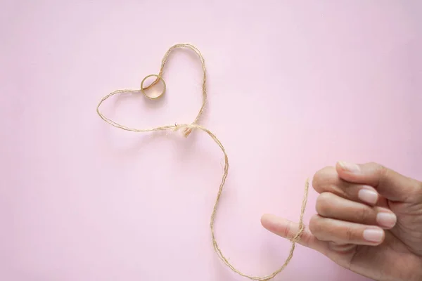 Herzförmiges Seil Und Goldring Mit Verschwommenem Frauenfinger Auf Rosa Papierhintergrund — Stockfoto