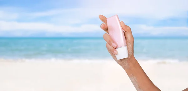 Hand woman holding sun block or sunscreen at coast with sand blue sea blue sky blured. sun cream lotion to protect UV. tourist summer travel holidays concept.