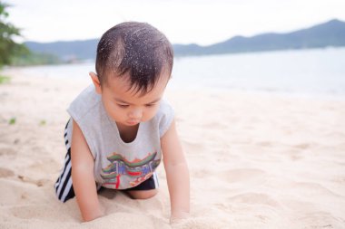 Küçük çocuk 2 yaşında tatlı bir çocuk sahilde kumsalda oturuyor bulanık bir okyanus doğası geçmişi var. Neşeli bir şekilde oynuyor. Turist ailesi yaz tatillerinde tropikal seyahatler.