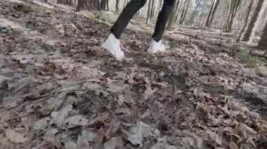 Beyaz spor ayakkabılarla ormanda düşen yaprakların arasından geçen kızın arka görüntüsü.