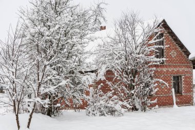 Ağaçların arkasında tuğla ev, çok kar, kışın köy.