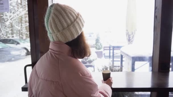 En flicka dricker kaffe i ett café med utsikt från fönstret på en snötäckt terrass, filmad bakifrån — Stockvideo