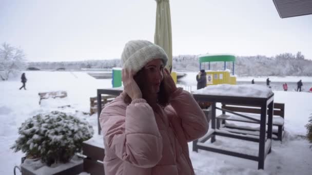 Een meisje in een roze donsjas en een wintermuts kijkt uit het raam op een winterterras in een café als in een spiegel — Stockvideo