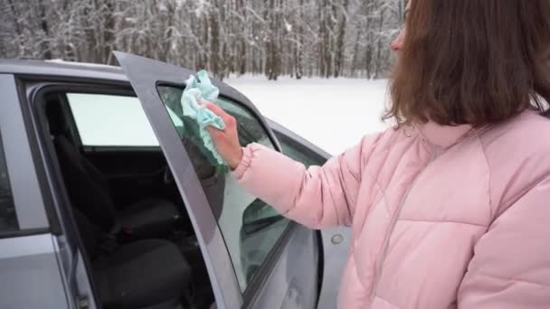 Menina em um inverno rosa jaqueta limpa a janela lateral de um carro com um pano no inverno, cuidados com o carro mão, encerando o vidro — Vídeo de Stock